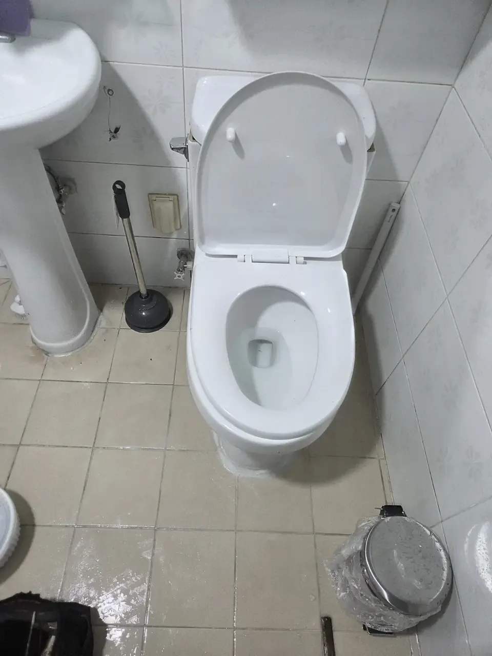 The same toilet refitted and clean, seat open, plunger standing beside it on a wet tiled floor