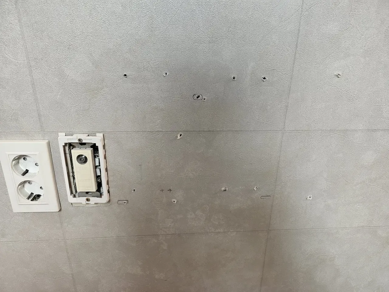 A tiled wall in an empty flat with around a dozen small drill holes above the power sockets