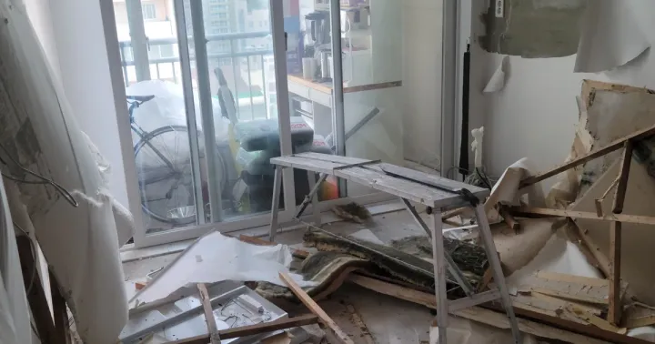 A living room with the entire ceiling torn out — battens, broken plasterboard and insulation piled on the floor after a leak from the flat above