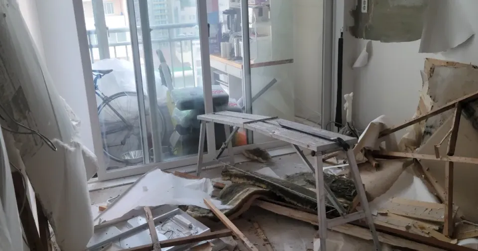 A living room with the entire ceiling torn out — battens, broken plasterboard and insulation piled on the floor after a leak from the flat above