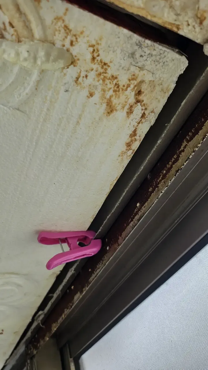 A close-up of a balcony window track with dark rust and grime built up along the aluminium frame beneath a peeling ceiling
