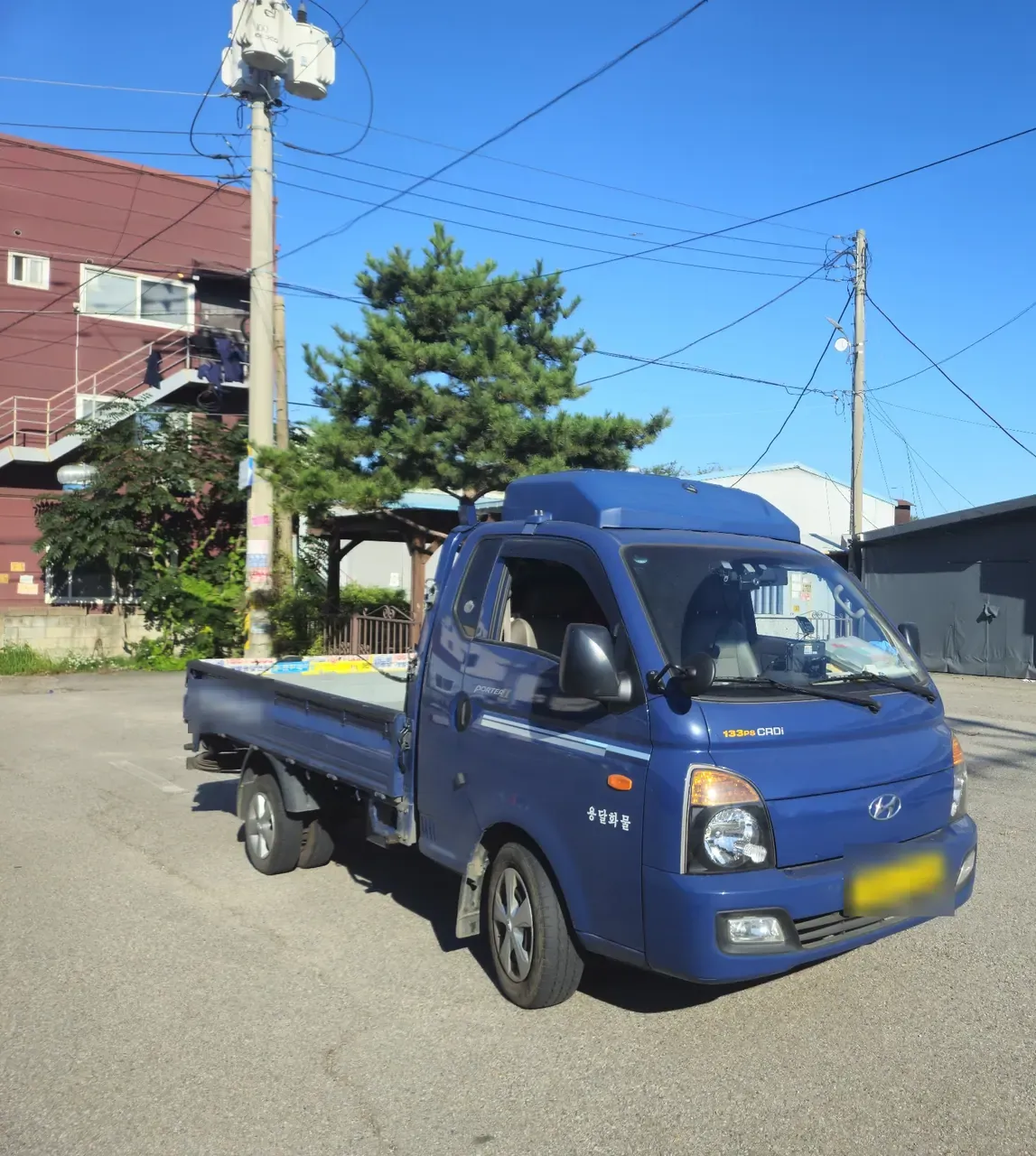 A blue Hyundai Porter 1-ton truck parked in a Korean back street, its cargo bed completely empty, with a power pole and a low-rise building behind